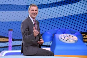 Beppe Convertini seduto su un tavolo e un telefono fisso gigante